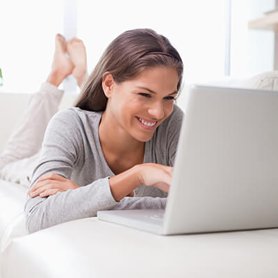 A smiling young woman lying on her sofa upside down while using her laptop