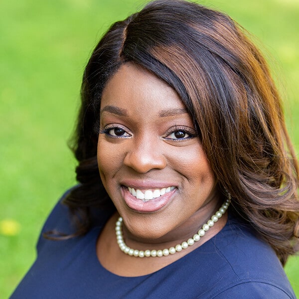 Dr. Ngozi Okoh smiling outside the dental office with a green background while wearing a pearl necklace